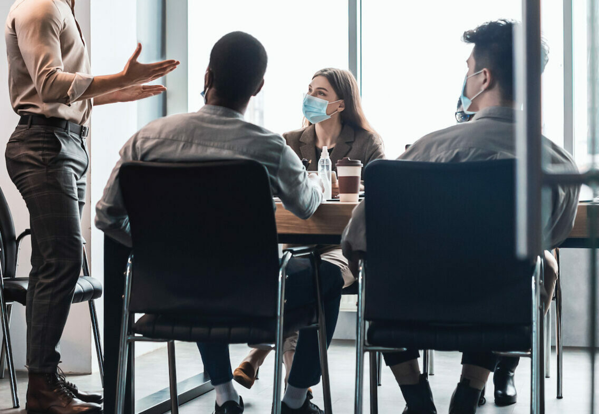 Modern Corporate Meeting Concept. Young Businessman In Medical Face Mask Giving Speech During Meeting With Coworkers In Office, Standing At Table In Board Room, Explaining Strategy And Management